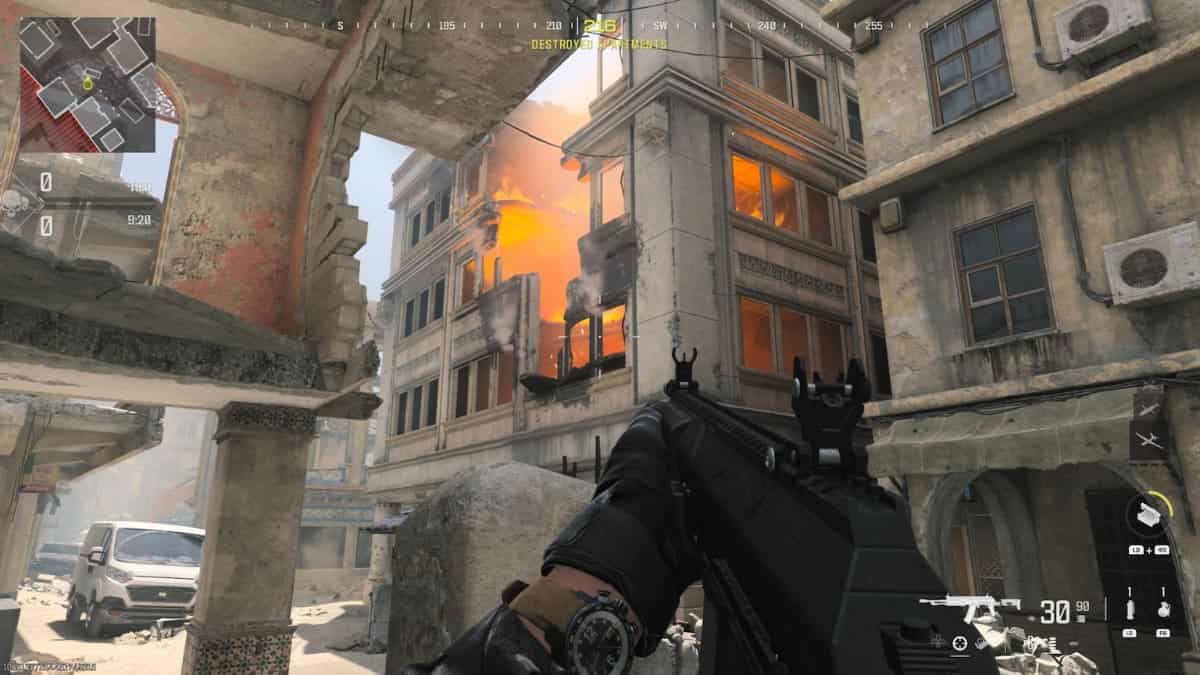 A first-person view from the MW3 Season 2 Reloaded video game shows an armed character with an assault rifle aiming towards a building engulfed in flames and smoke.
